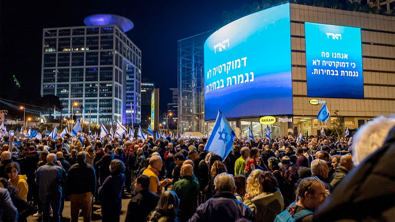 Demonstranter mot den israeliska regeringen samlas under en reklamskylt för Haaretz, med budskapet ”demokratin upphör inte med valet”. Foto: Oren Rozen/Wikimedia.